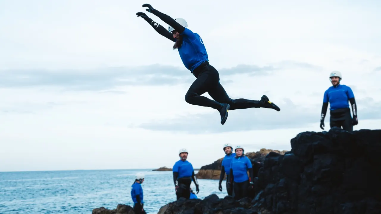 Person in blue jacket jumping off rocky cliff into sea whilst group in wetsuits watches below.