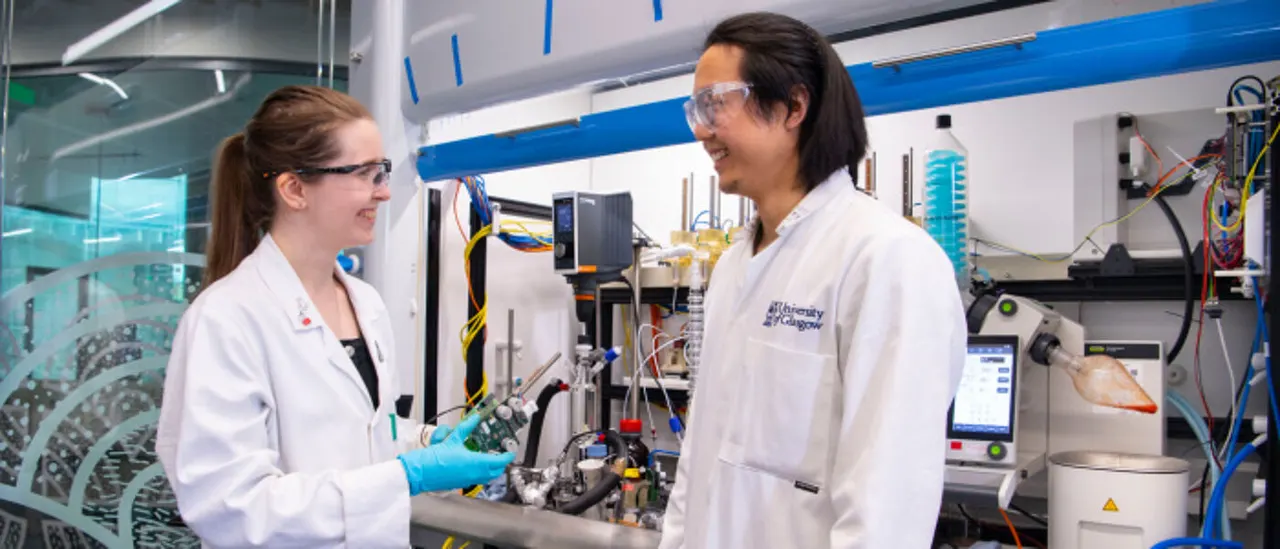 Two researchers in white lab coats and safety glasses work together at scientific equipment in a modern laboratory.