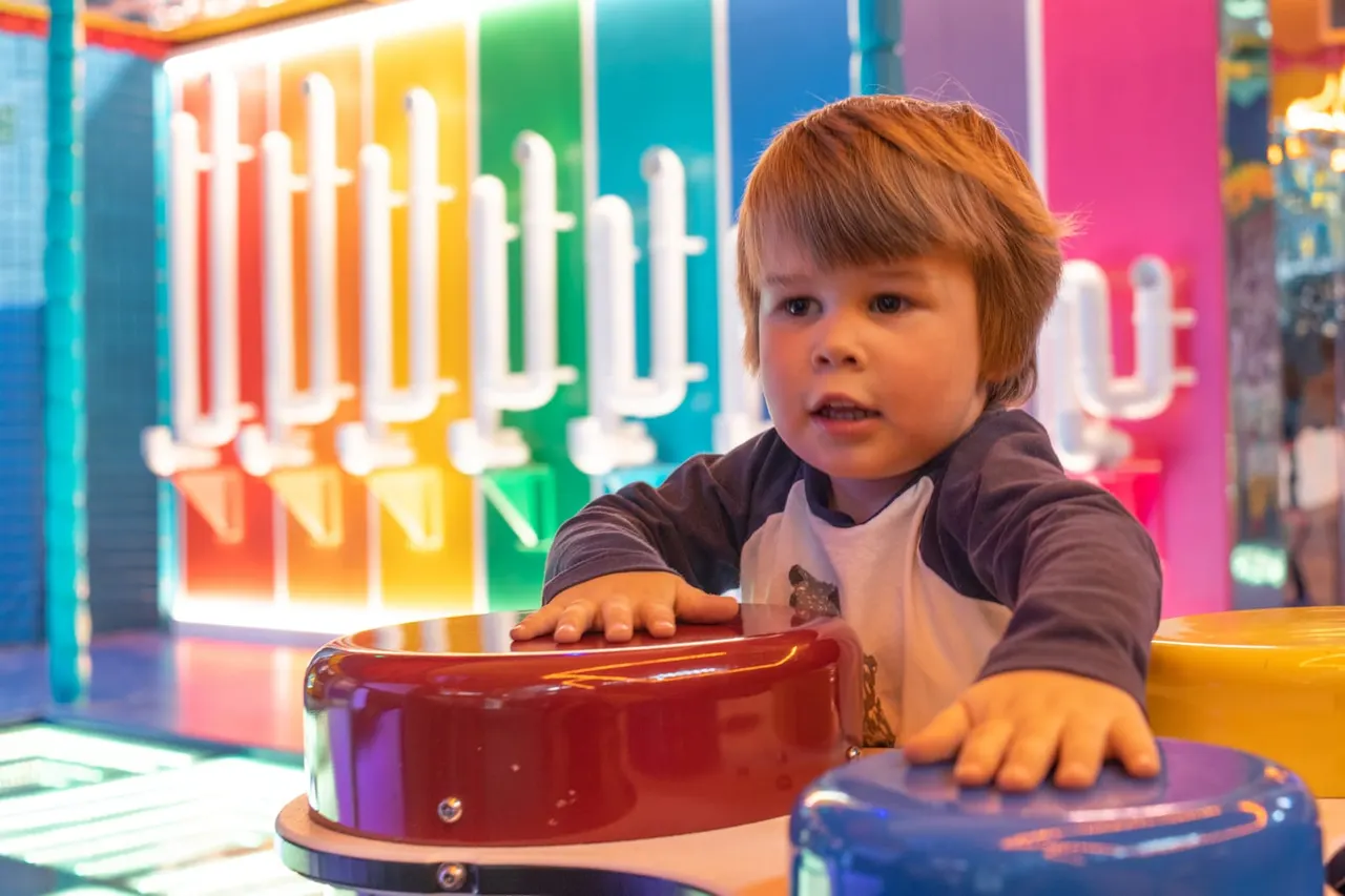 Young child playing with a red drum at a colourful soft play venue with vibrant rainbow-lit panels in the background.