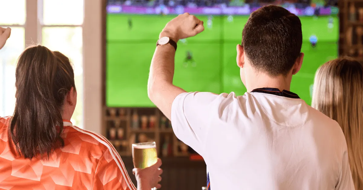 Group of people watching football match on large screen whilst holding drinks in a bar.
