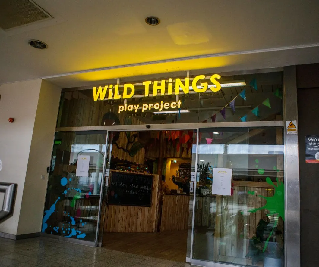 Exterior storefront of Wild Things play project with yellow neon signage, glass doors and windows decorated with colourful animal graphics.