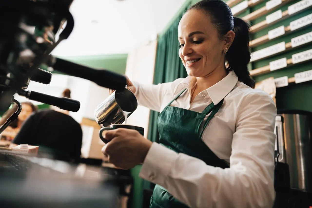 Barista in green apron pouring milk into a cup whilst making a coffee drink behind the counter.