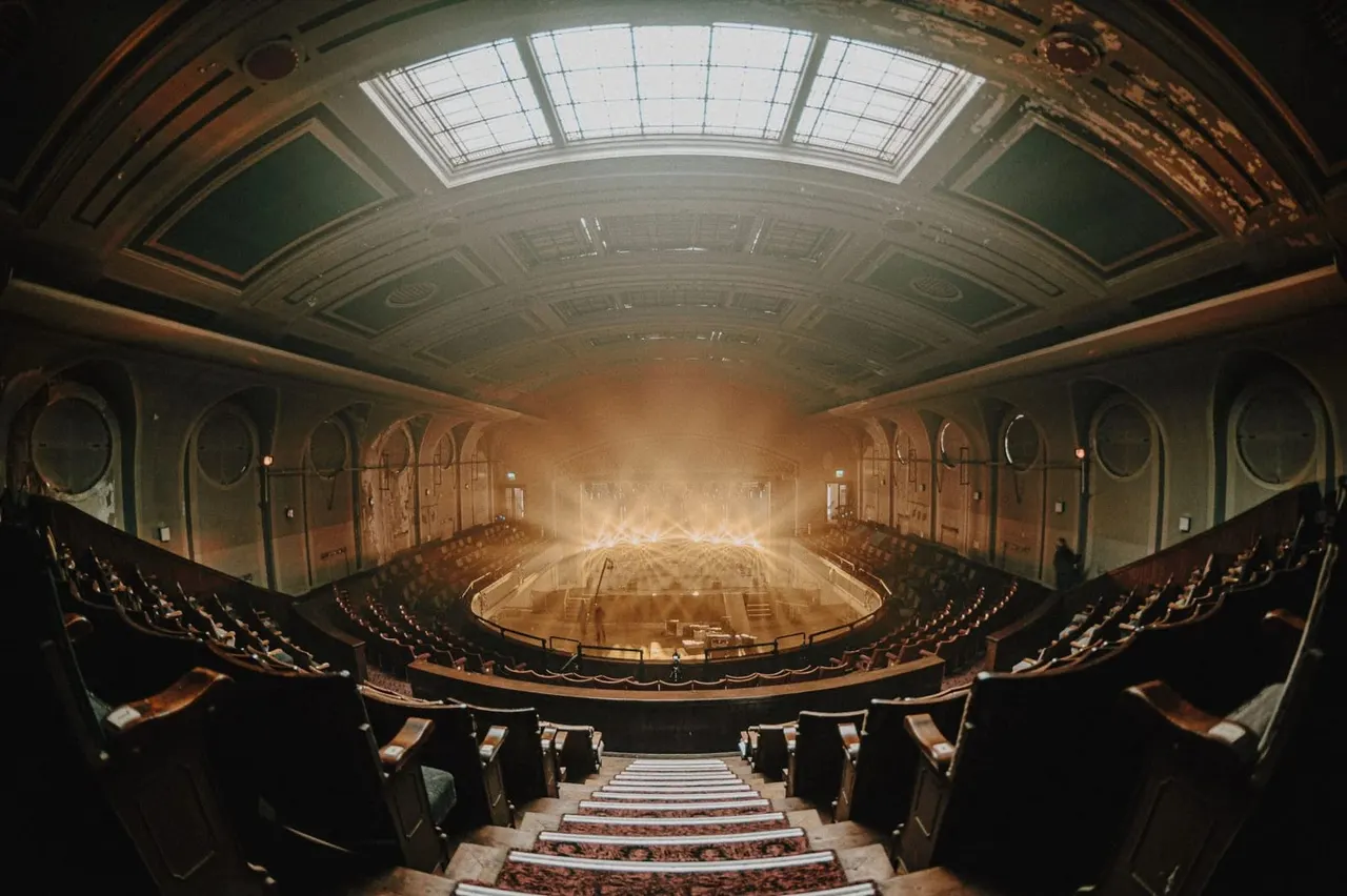 Interior of a historic theatre auditorium with tiered seating, ornate ceiling with skylight, and illuminated stage.