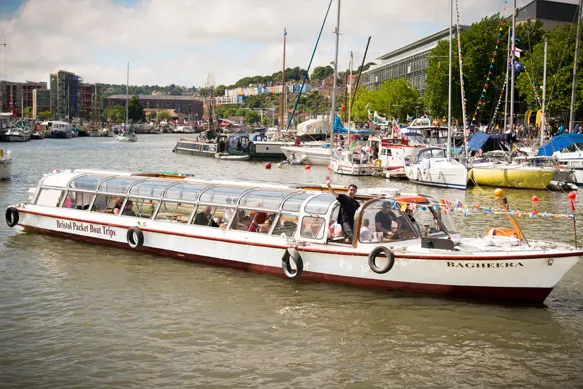 A long passenger boat moored in a busy harbour with sailing yachts and waterside buildings in the background.