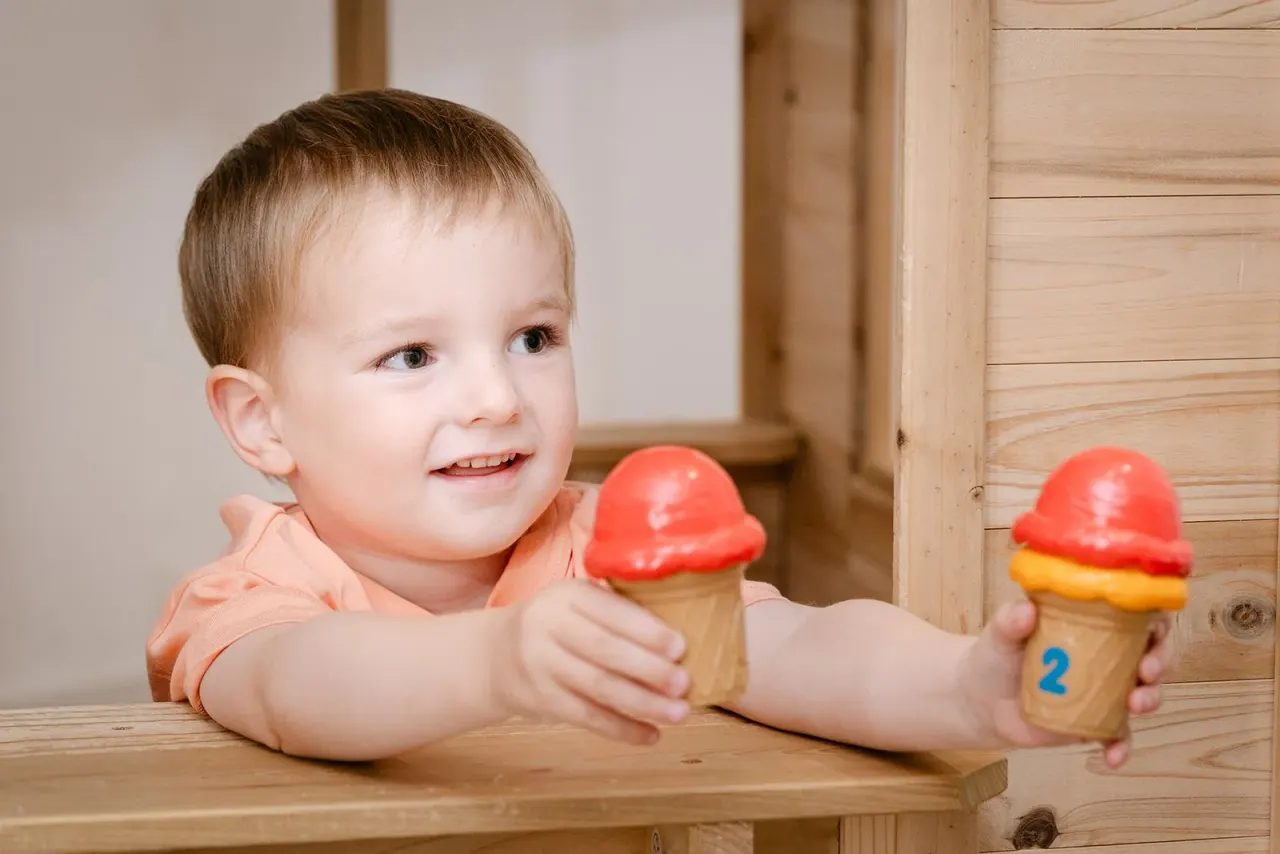 A young child in an orange shirt plays with toy ice cream cones with red tops at a wooden table.