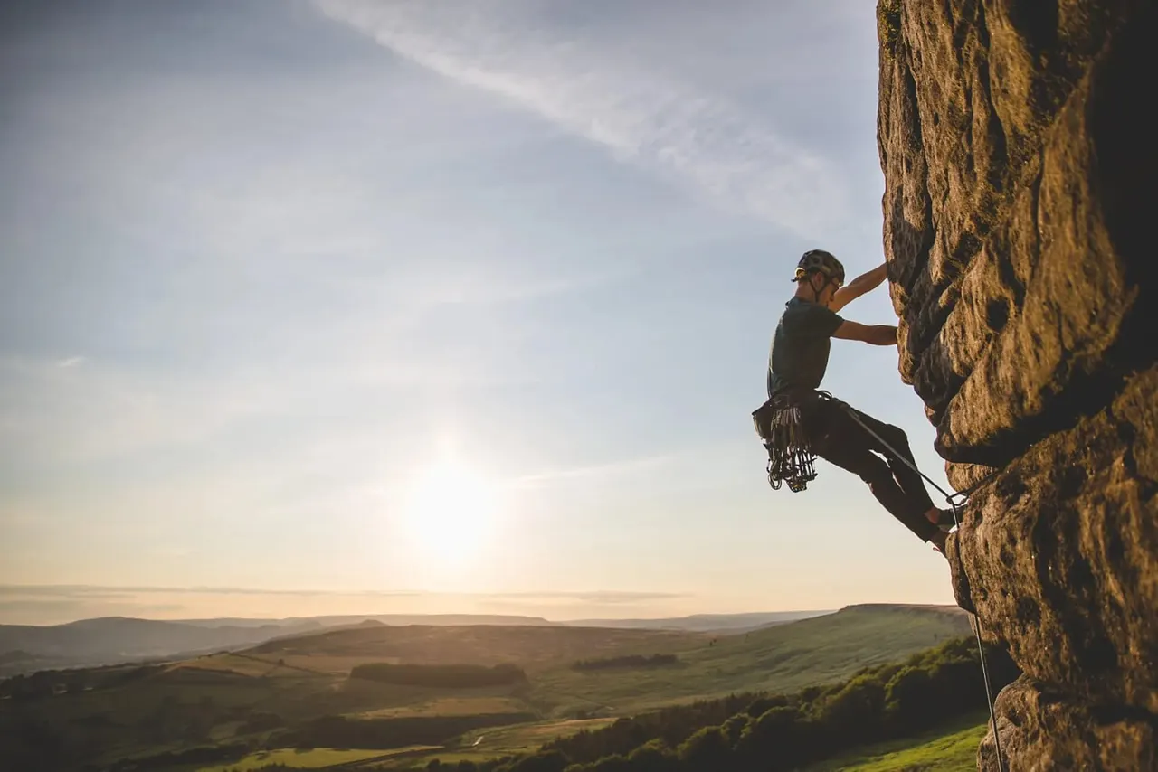 Climber ascending a sandstone cliff face with expansive rolling countryside visible below.
