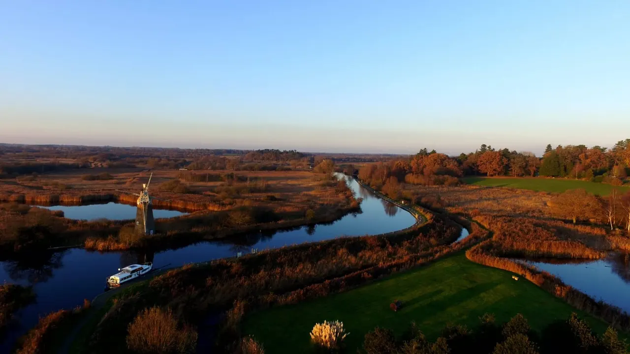 Aerial view of a windmill and boat on a winding river through autumn wetland and woodland landscape.