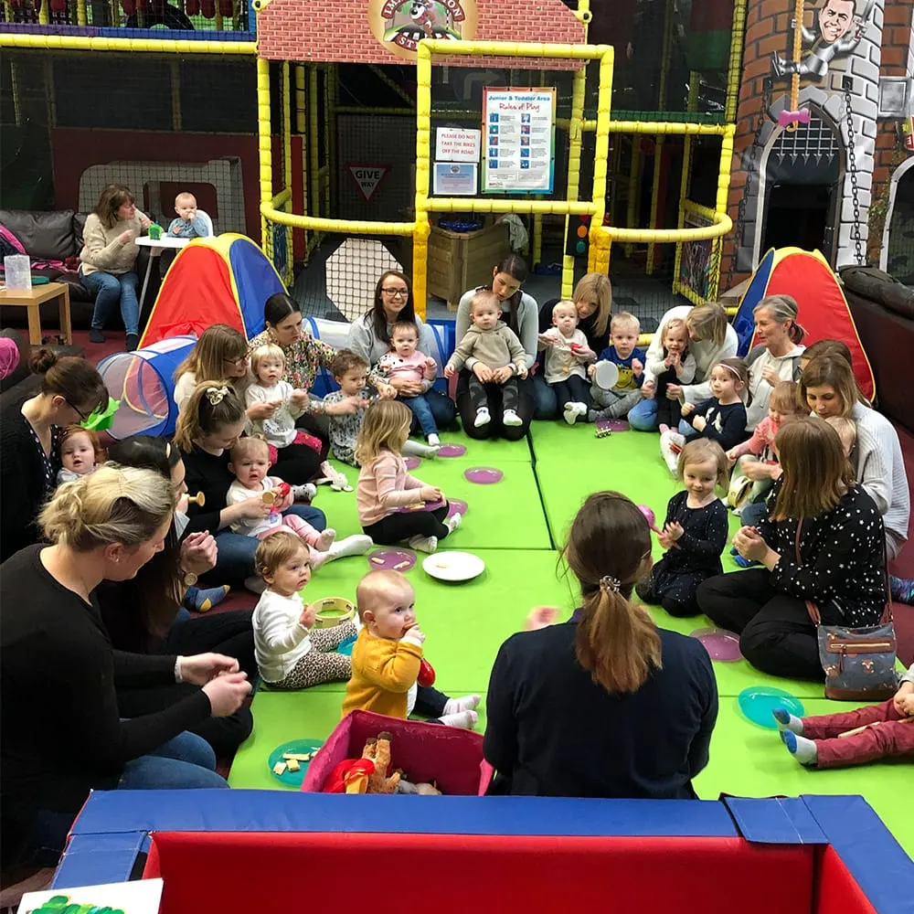 Adults and toddlers sitting together on a bright green padded floor in an indoor play facility with coloured tunnels and climbing structures.