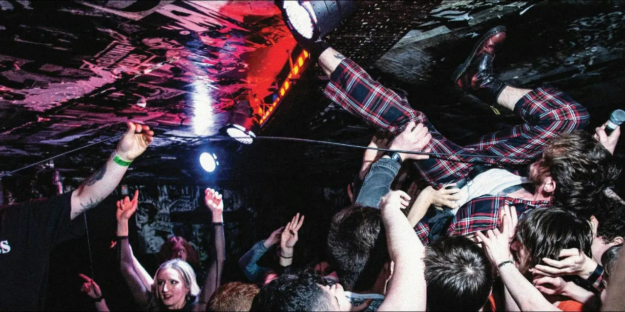 Crowded nightclub dance floor with people crowd-surfing and dancing under colourful red and pink lighting.