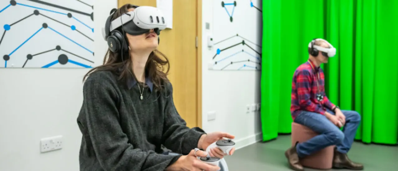 Woman wearing a VR headset and holding controllers whilst another person in a VR headset watches from the side.