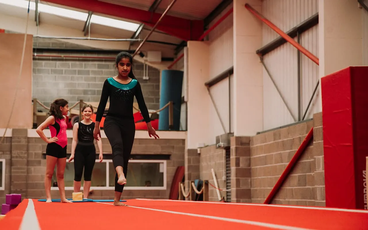 An instructor leads two young girls on a red gymnastics mat in an indoor sports facility.