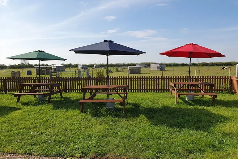 Picnic tables with green, navy and red umbrellas arranged in a grassy outdoor seating area with wooden fencing and farmland beyond.