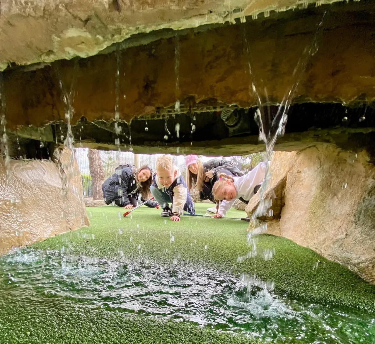 Children playing mini golf under a stone bridge with water fountains flowing overhead.