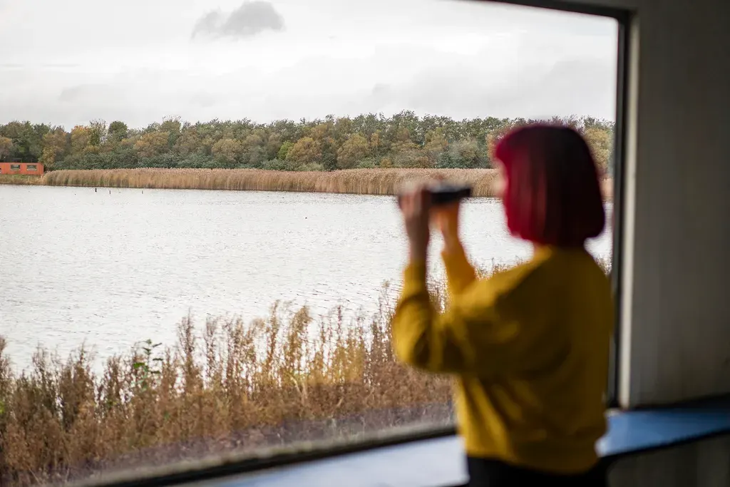 Person in yellow jacket with binoculars looking out at lake, reeds and woodland from indoor viewing area.
