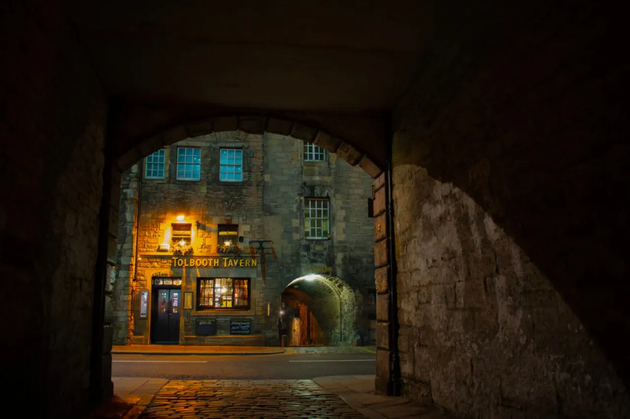 Historic stone tavern building viewed through a narrow archway at night, with warm lighting illuminating its facade and blue-lit windows.