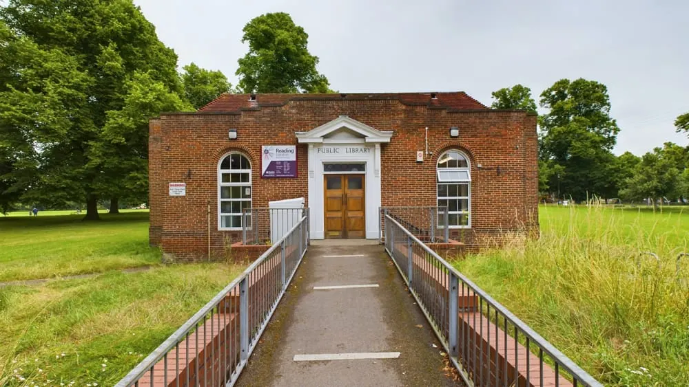 A brick public library building with wooden doors and arched windows, approached via a paved pathway with metal railings, set in a grassy park surrounded by trees.
