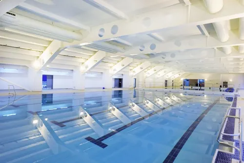 Indoor swimming pool with multiple lanes separated by lane dividers, bright white ceiling with skylights, and empty poolside.