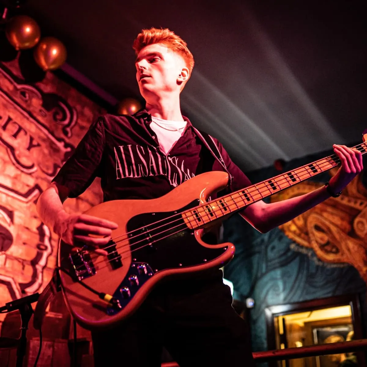 Musician playing bass guitar on stage under red lighting with decorative backdrop.