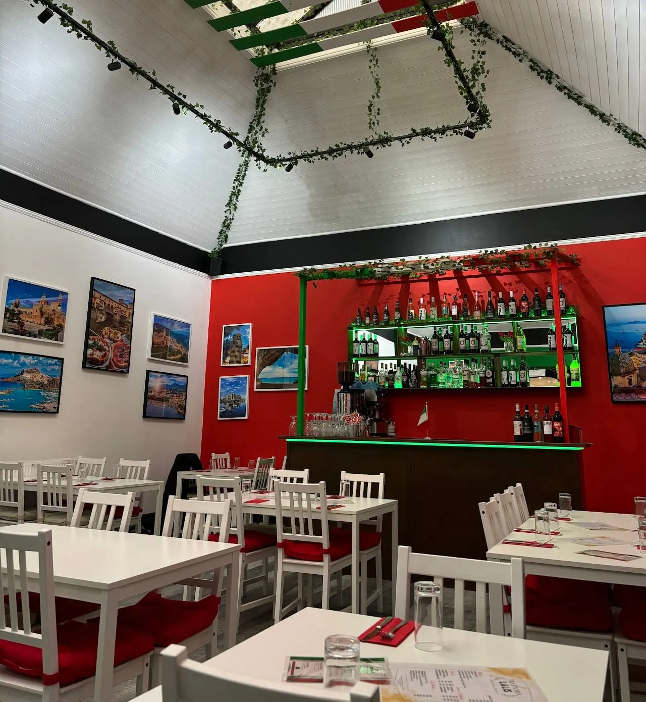 Restaurant interior with white tables and red-seated chairs, red accent wall with bar, hanging greenery, and coastal photographs.