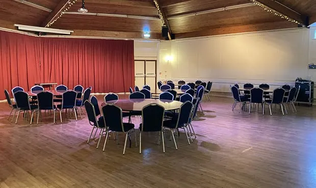 A spacious theatre hall with round tables and chairs arranged for dining or events, red curtains, and wooden beams.