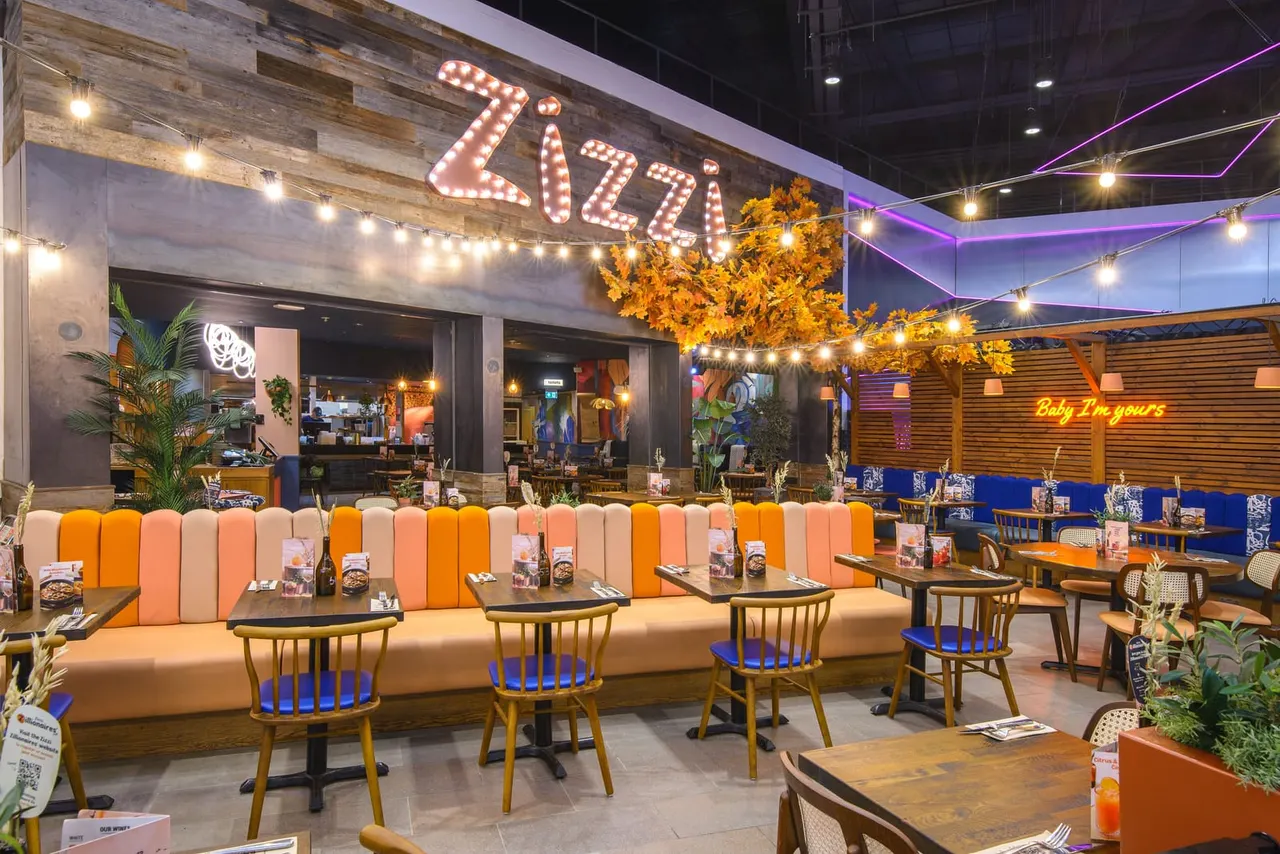 Modern restaurant interior with colourful booth seating, wooden chairs, and a large illuminated sign reading "Zizzi" above the dining area.