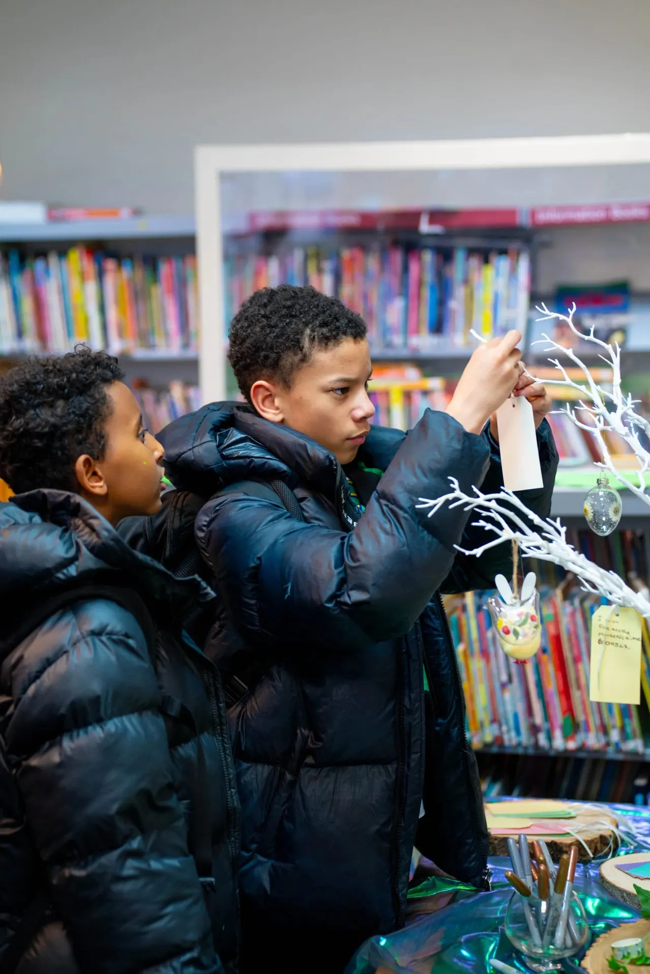 Two children in black jackets decorating a white branch with ornaments in a library with bookshelves behind them.