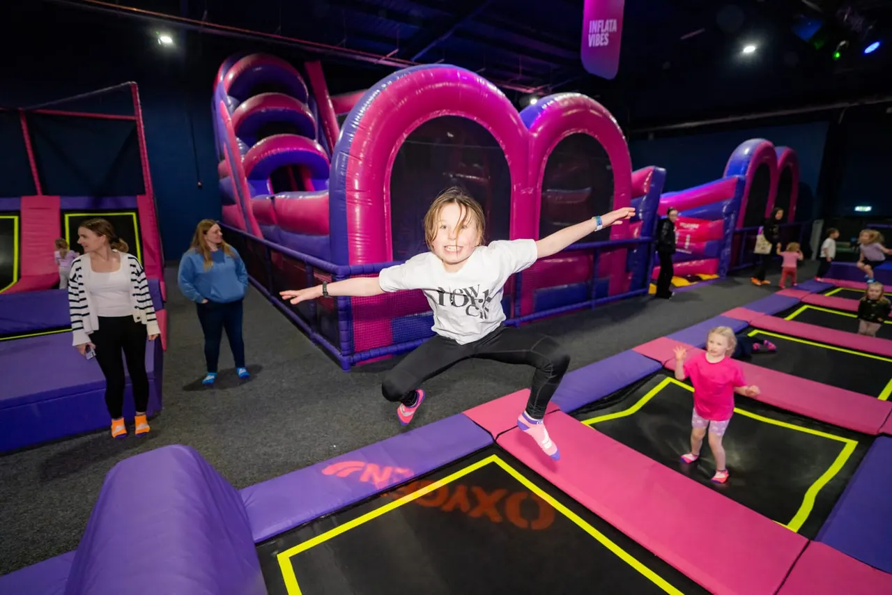 Child bouncing on a trampoline with arms outstretched in an indoor play arena filled with pink and purple inflatable structures.