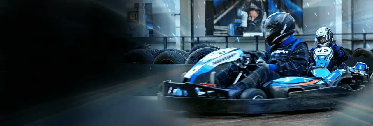 Two people in helmets racing blue and black go-karts on an indoor track with tyres and barriers visible.