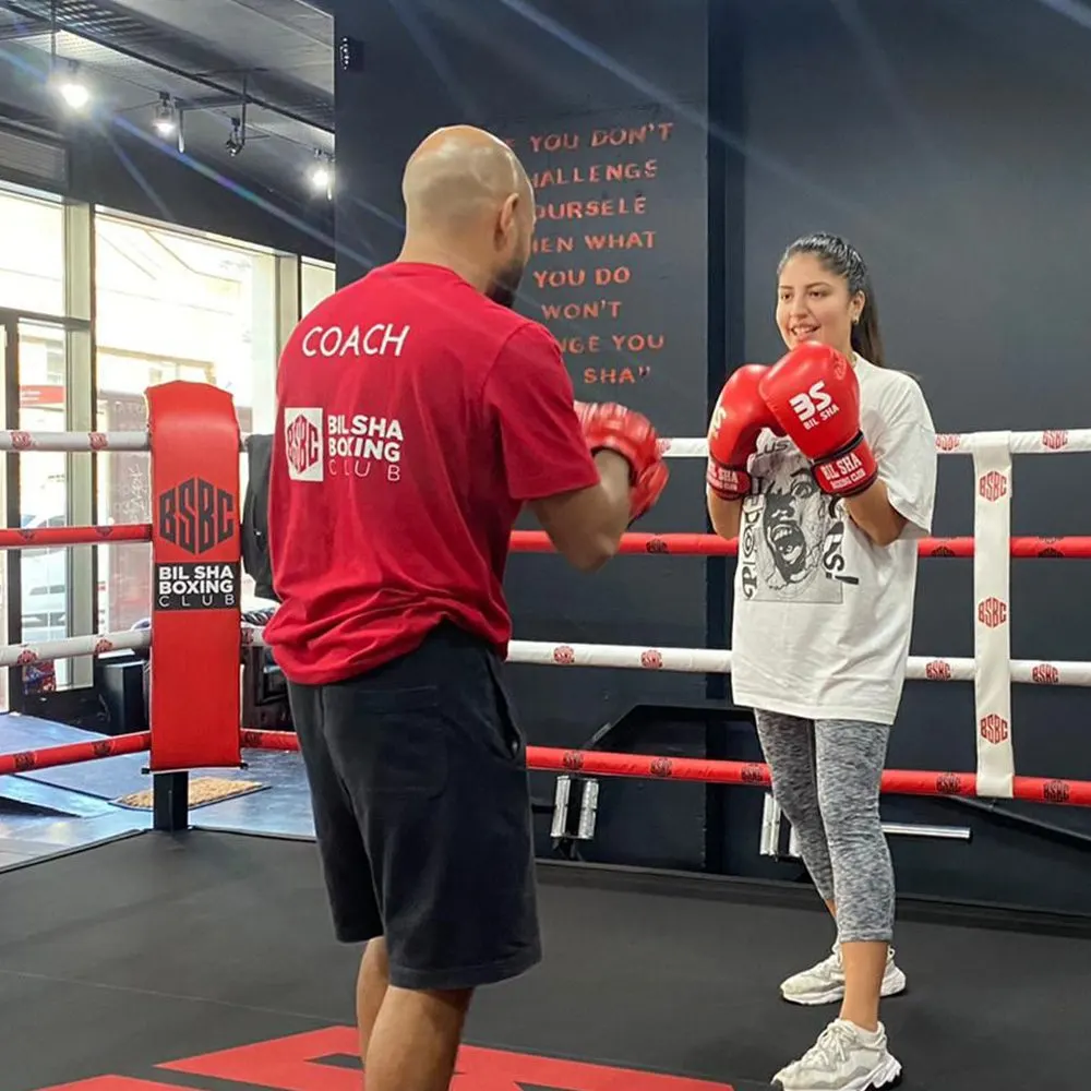 A coach in a red shirt faces a young woman wearing red boxing gloves inside a boxing ring at a gym.