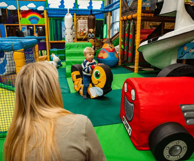 Toddler playing on a ride-on toy in a colourful indoor play area with an adult watching nearby.