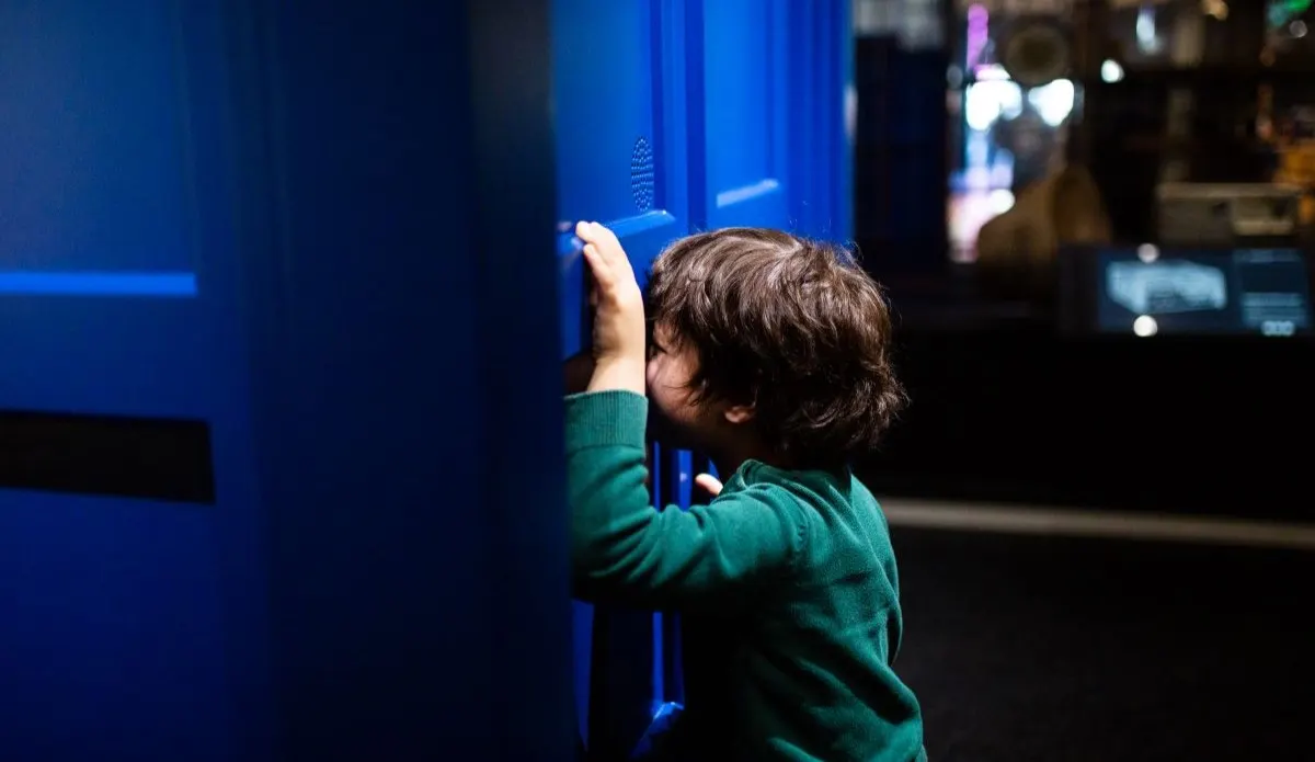 A young child in a green jumper interacts with a blue illuminated touchscreen display in a museum setting.