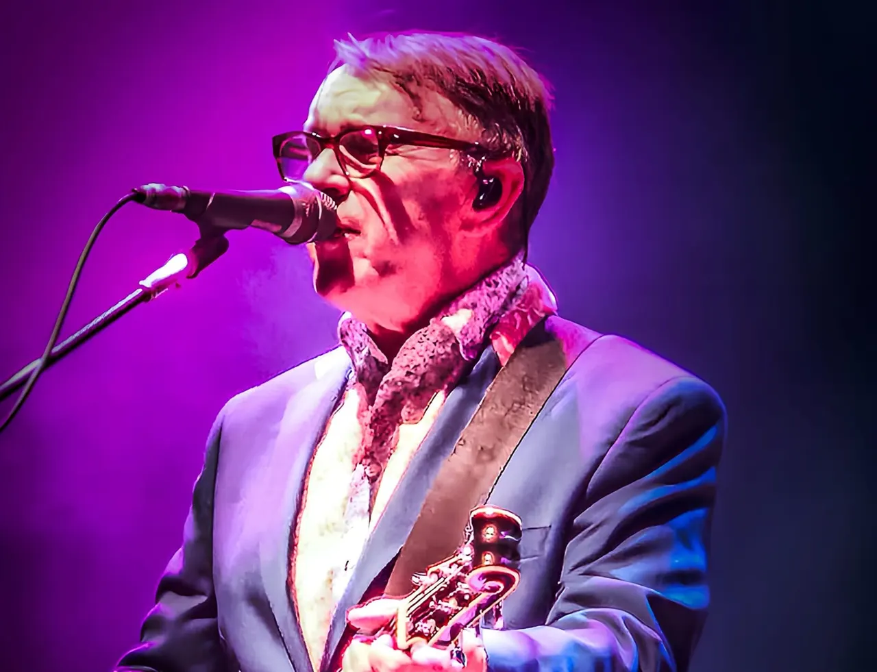 Performer wearing glasses and suit singing at microphone under purple stage lighting.
