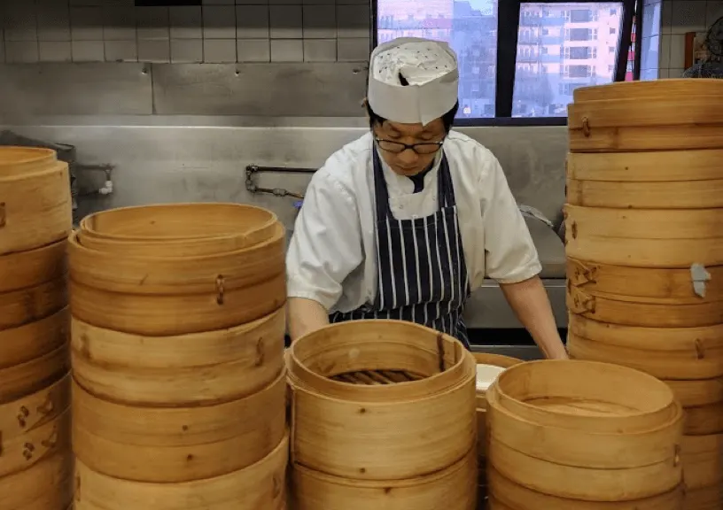 Chef in white hat and striped apron working with bamboo steamers in a commercial kitchen.