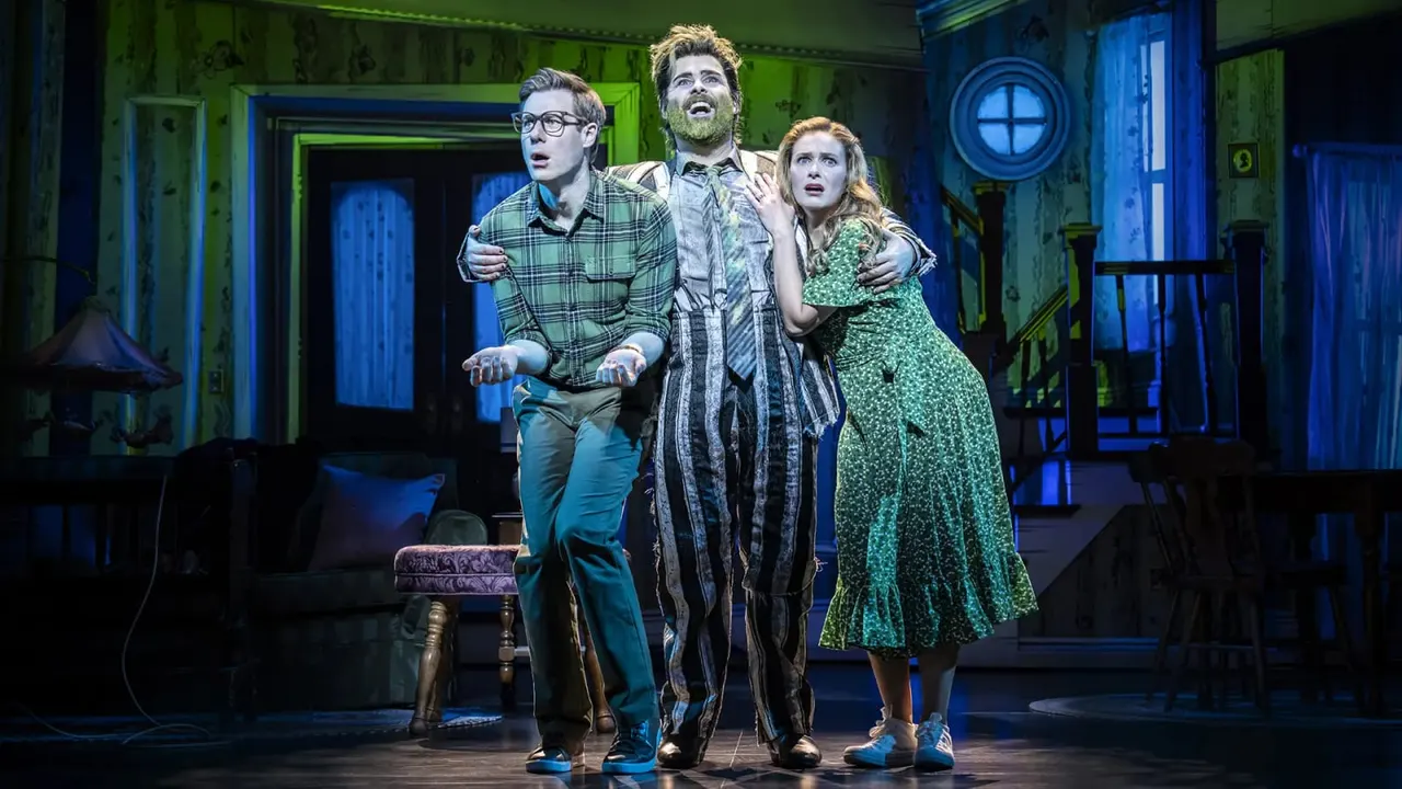 Three actors perform on stage in a stylised green and blue interior set, with a man in the centre with his arms around two younger performers on either side.