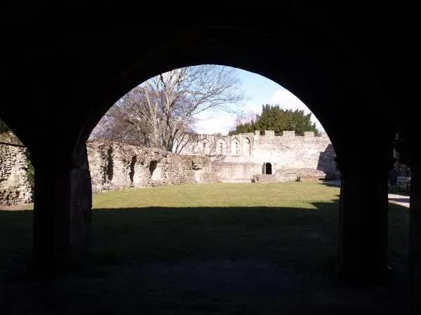 Medieval ruins viewed through a dark stone archway, with bare trees and grassed courtyard beyond.