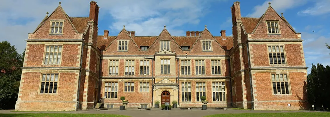 A large red-brick Victorian mansion with symmetrical gabled wings and manicured lawns under a partly cloudy sky.