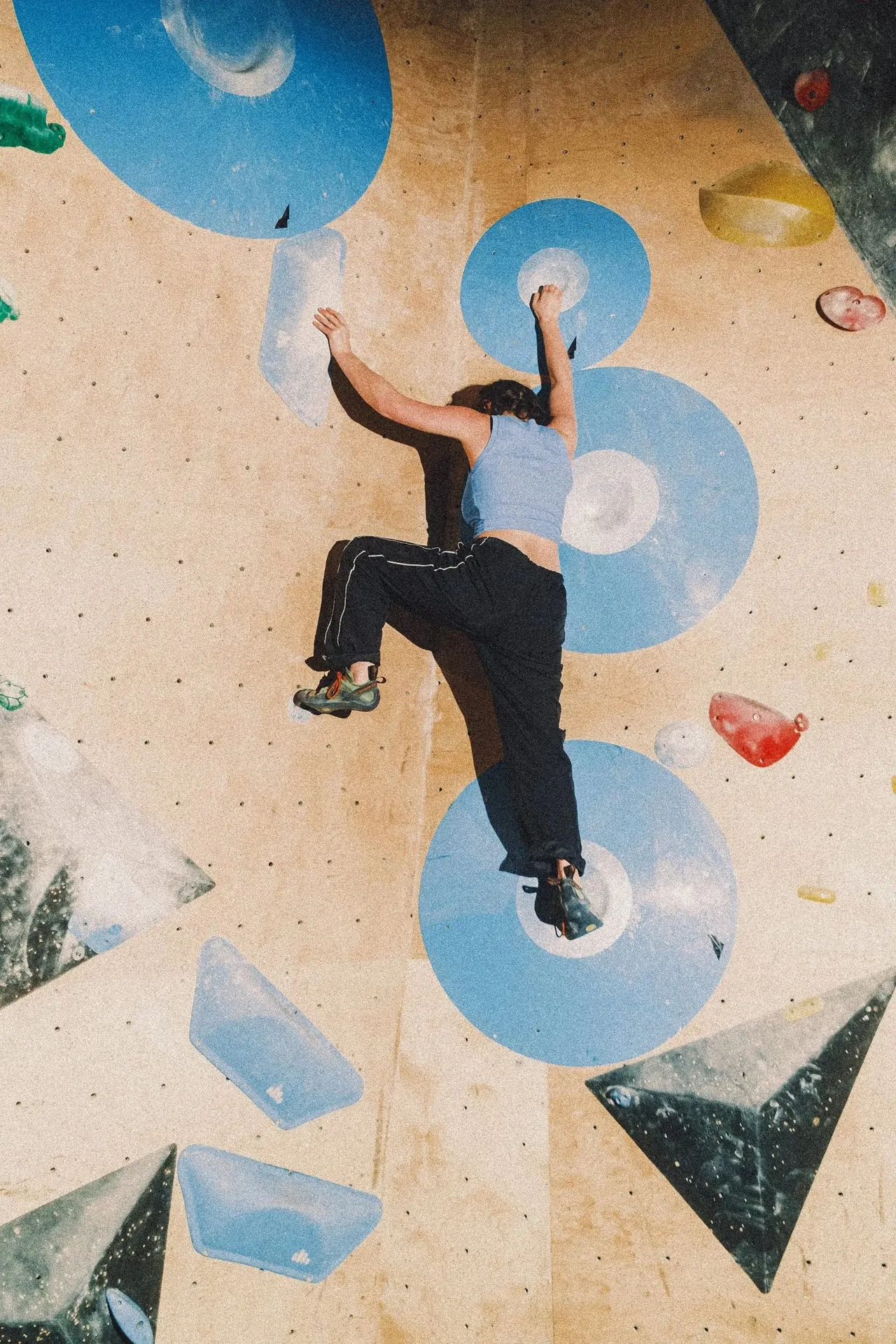 Person climbing an indoor rock wall with blue circular and triangular holds, viewed from above.