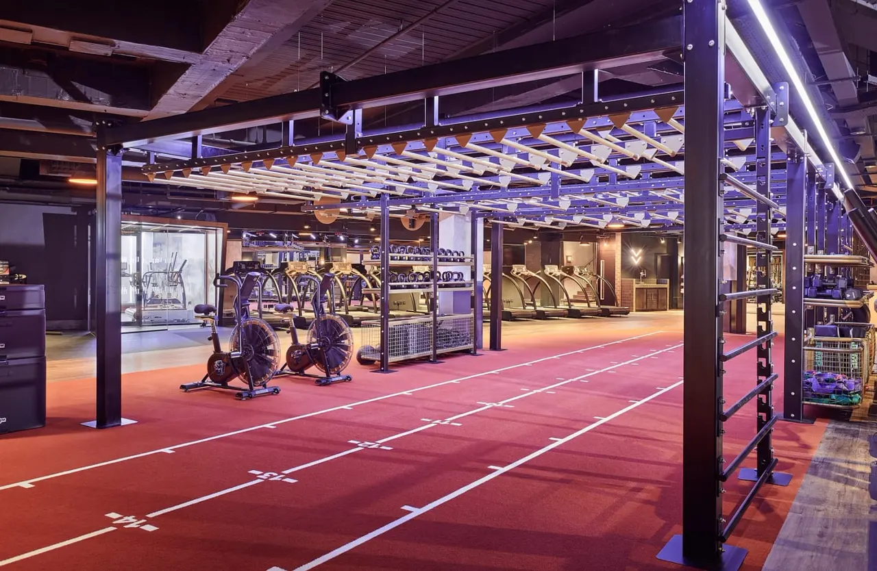 Modern gym interior with exercise bikes, treadmills and fitness equipment on red flooring under purple lighting.