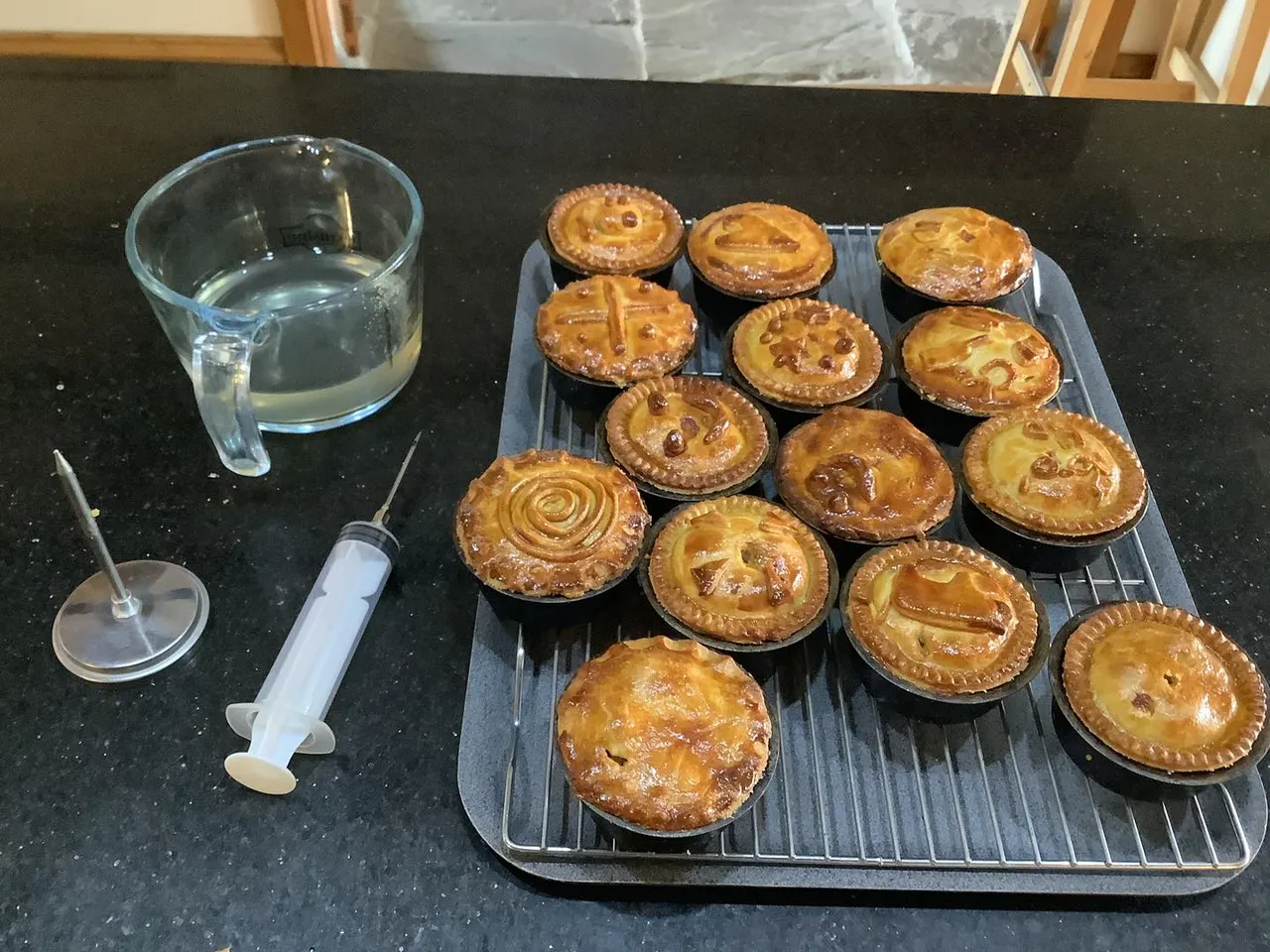 Freshly baked individual meat pies cooling on a rack with a glass of water and piping syringe on a kitchen worktop.