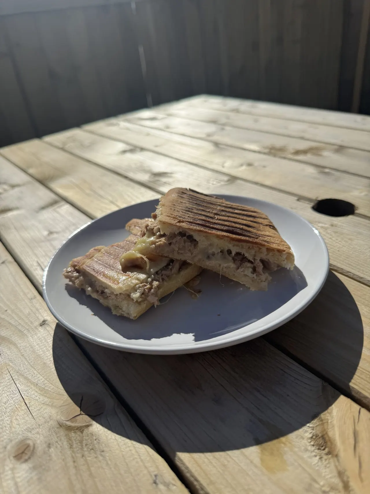 Toasted sandwich with filling on a white plate sat on a wooden table in sunlight.