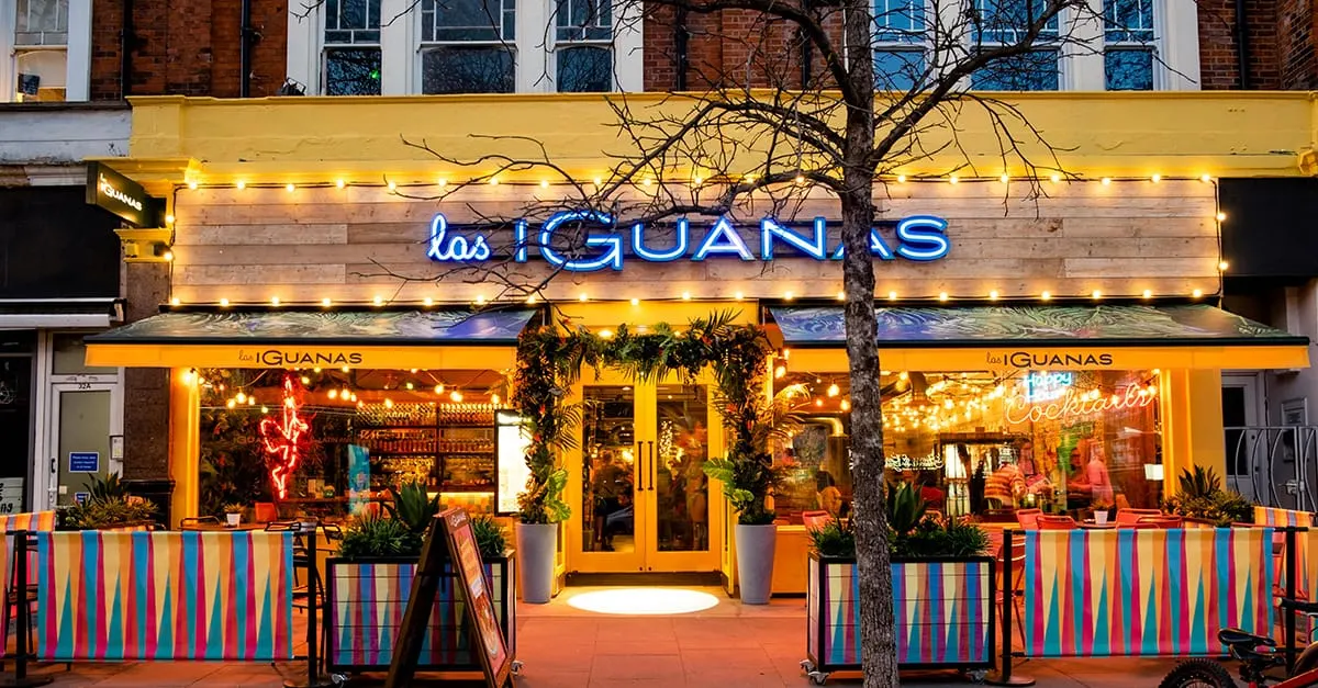 Brightly lit restaurant frontage with blue neon signage, yellow frame, colourful striped seating barriers, and potted plants at dusk.