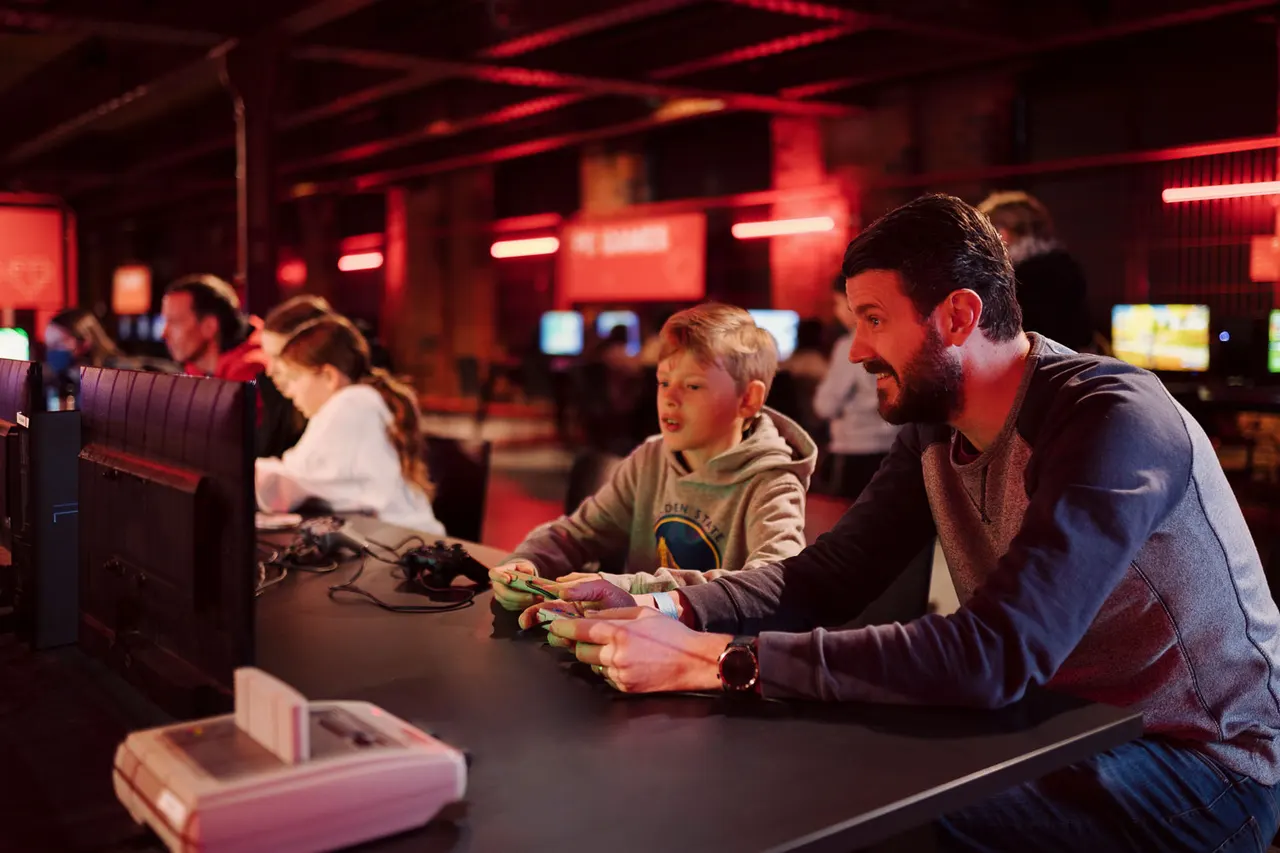 Adult and child playing video games together in a red neon-lit arcade room with other visitors in background.