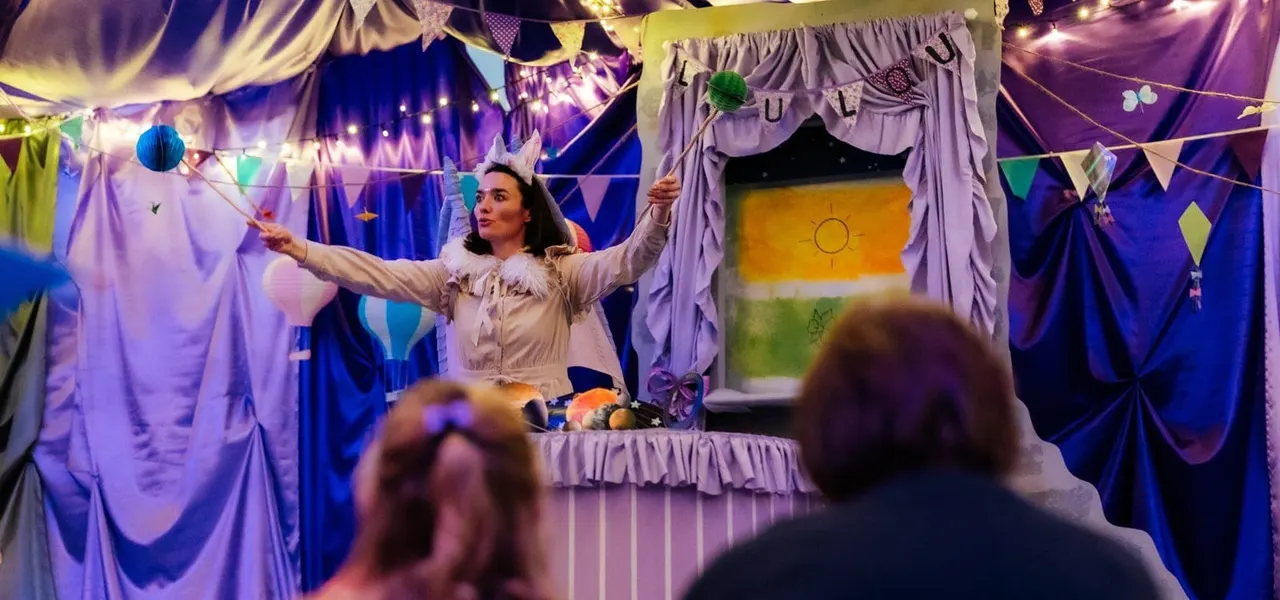 A performer entertains children in a colourful fabric-draped theatrical tent decorated with bunting and fairy lights.