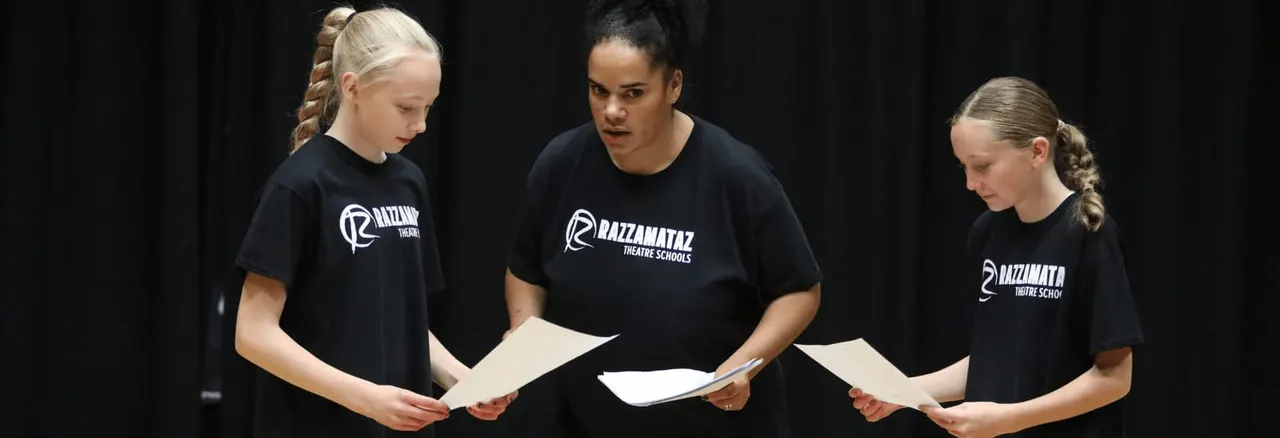 Three young performers wearing Razzamataz Theatre Schools t-shirts hold scripts during a rehearsal on stage.