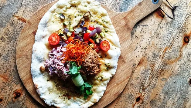 Flatbread topped with shredded meat, red cabbage, tomato slices and green herbs on a wooden board.