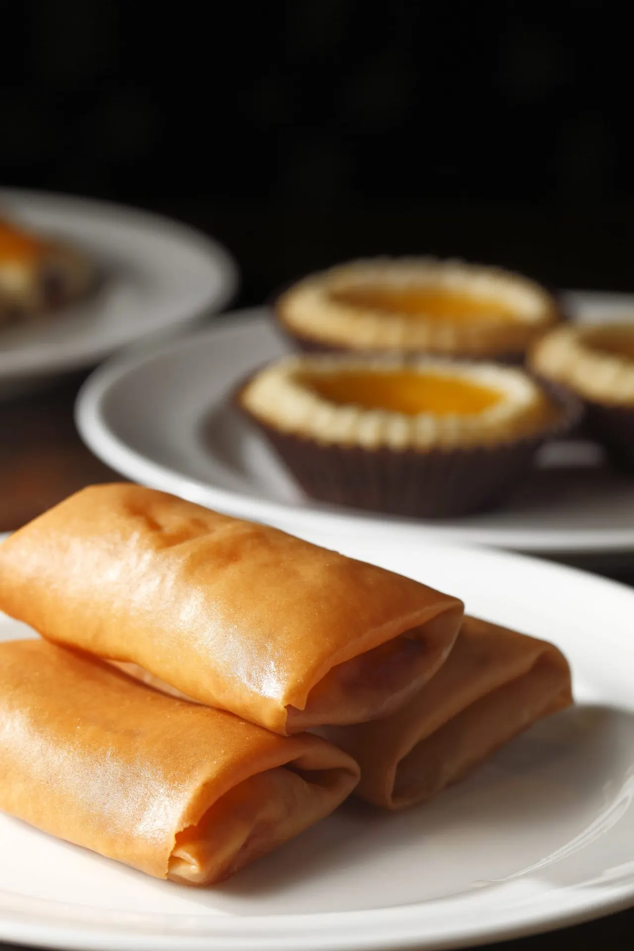 Two golden spring rolls on a white plate with pastry tarts blurred in the background.