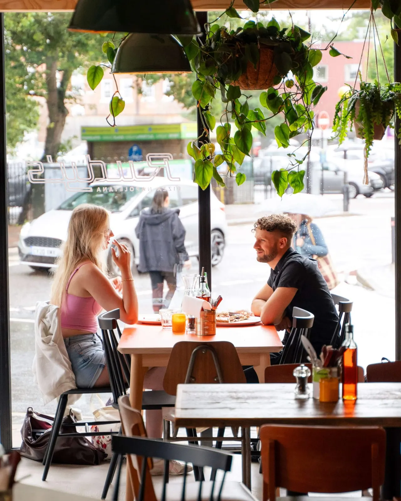 Two people dining at a table by the window of a restaurant with hanging plants and street view beyond.