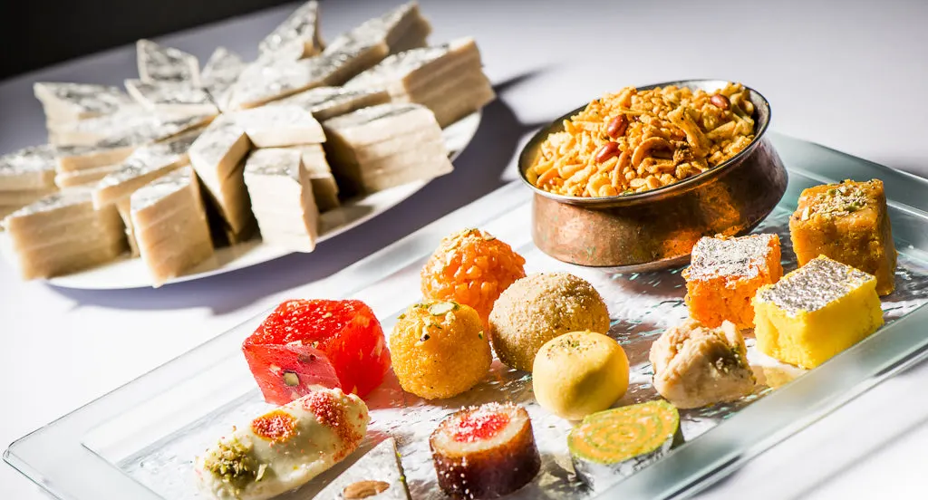 A platter of Indian sweets including gulab jamun, barfi and other confections, with a bowl of mixture snack and sliced cheese on the side.