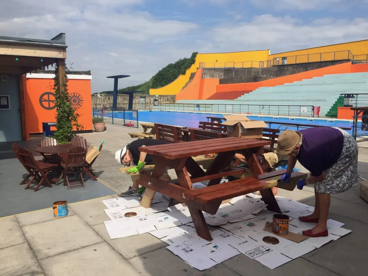 Two people painting a wooden picnic table at an outdoor leisure venue with a colourful pool building and seating areas in the background.
