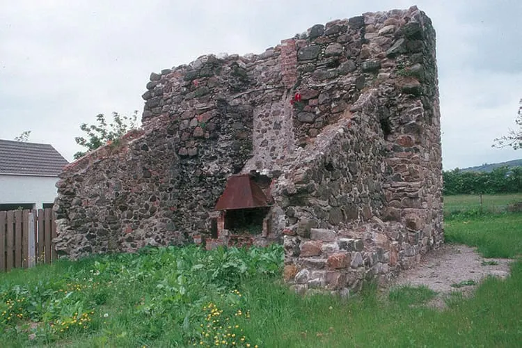 Ruined stone structure with a dark doorway opening, surrounded by overgrown grass and wildflowers in a rural landscape.